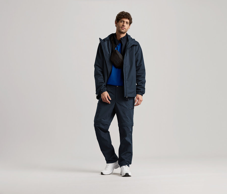 Muž stojí v tmavě modré bundě, kalhotách a bílých teniskách, s černou taškou přes rameno.