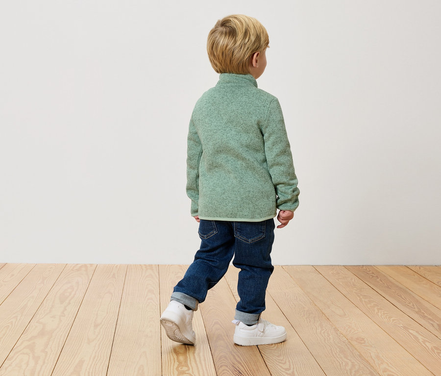 Dítě kráčí zády ke kameře v zeleném fleecovém svetru, džínách a bílých teniskách na dřevěné podlaze.