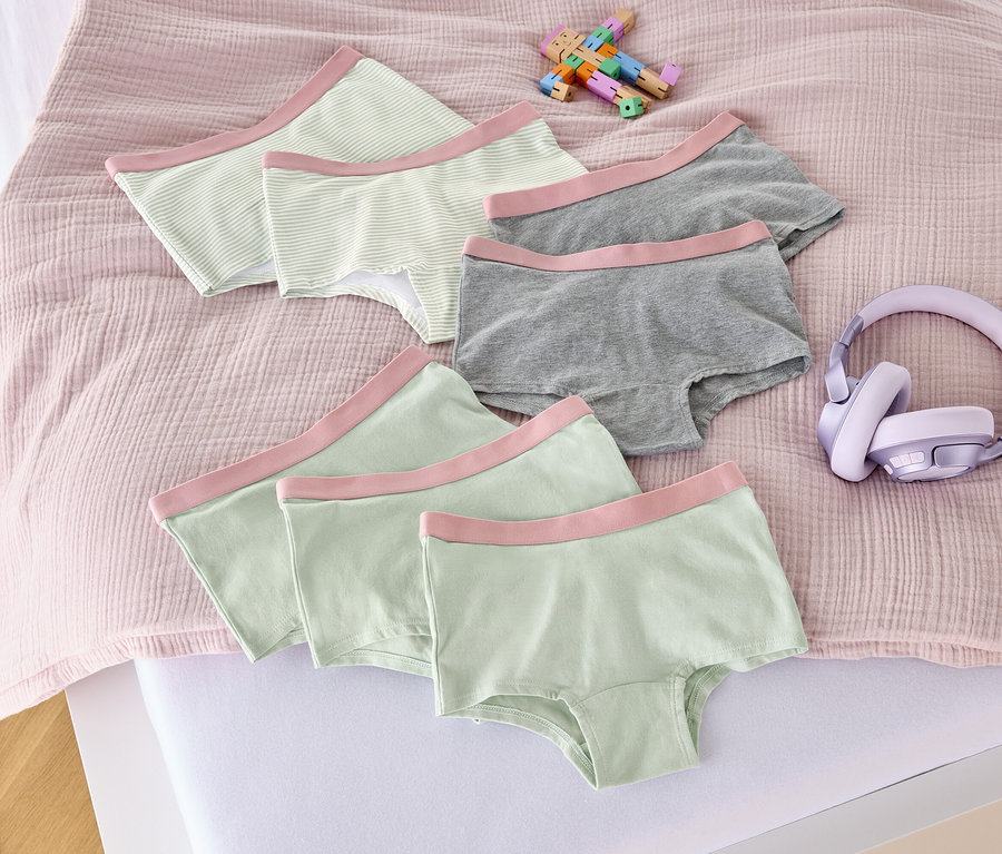 Na posteli leží sedm dívčích kalhotek v starorůžové barvě, hračka a sluchátka.