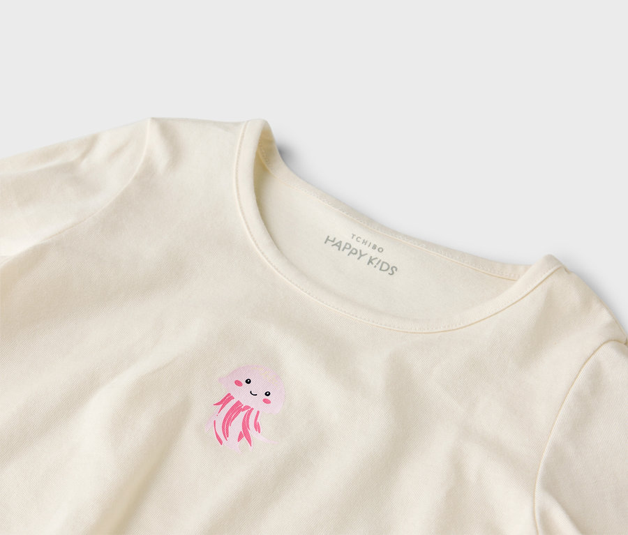 Detailní záběr na krémové tričko s potiskem růžové medúzy a potiskem "Tchibo Happy Kids" na krku.