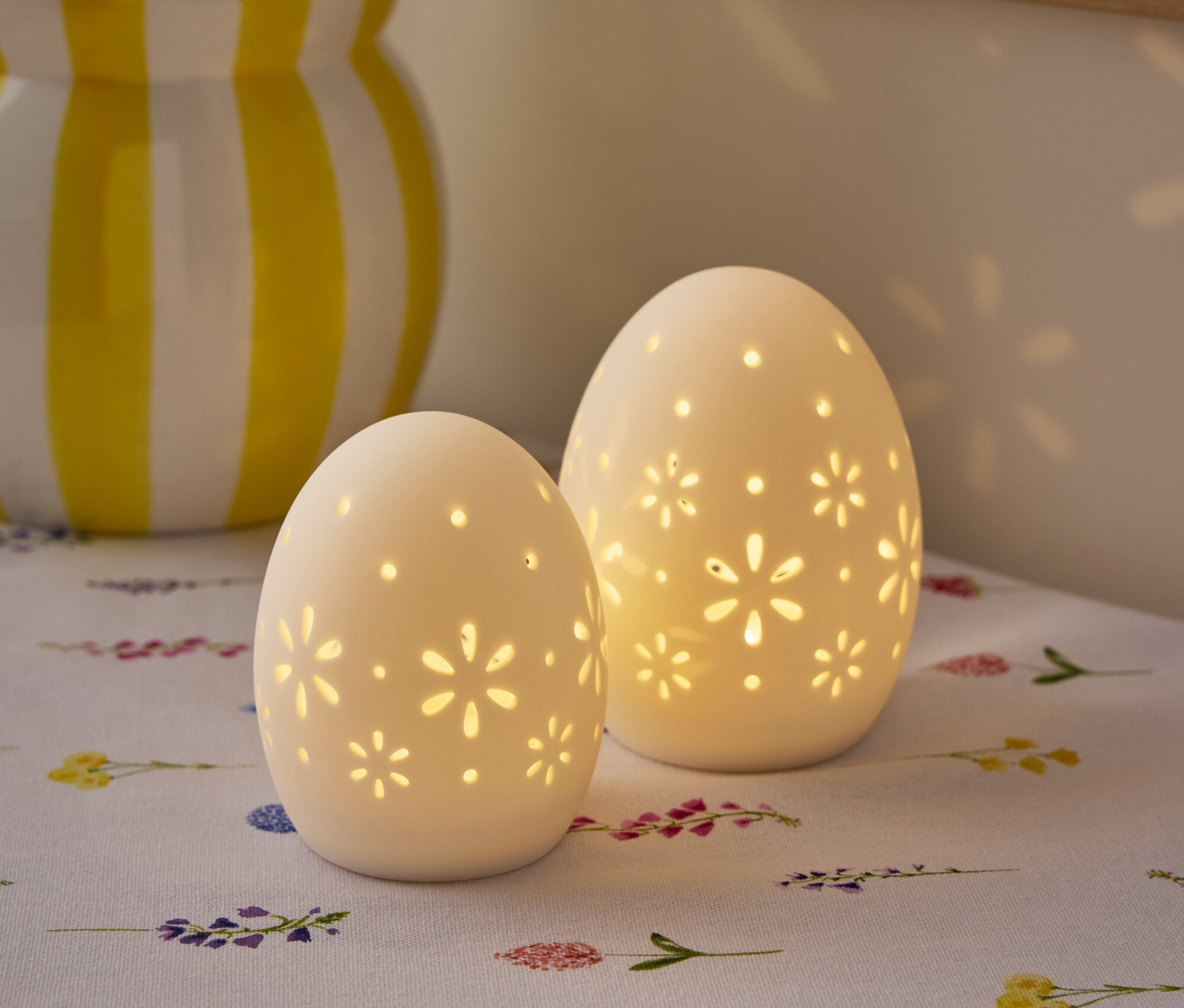 Dvě bílé lampy ve tvaru vejce s květinovým vzorem stojí na stole s vázou květin v pozadí.