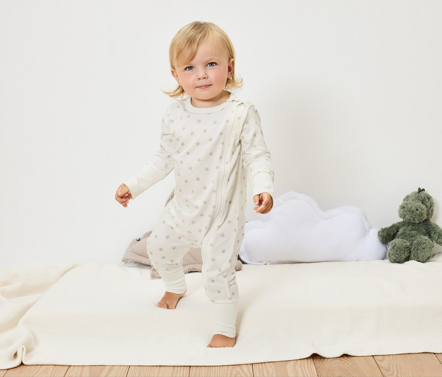 Blonďaté batole v bílém overalu s hvězdami stojí bosé na bílé dece vedle obláčkového polštáře a zelené plyšové hračky.