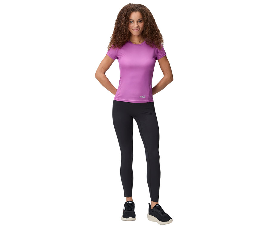 Žena stojí s rukama za zády, má na sobě fialové tričko, černé legíny a tenisky.
