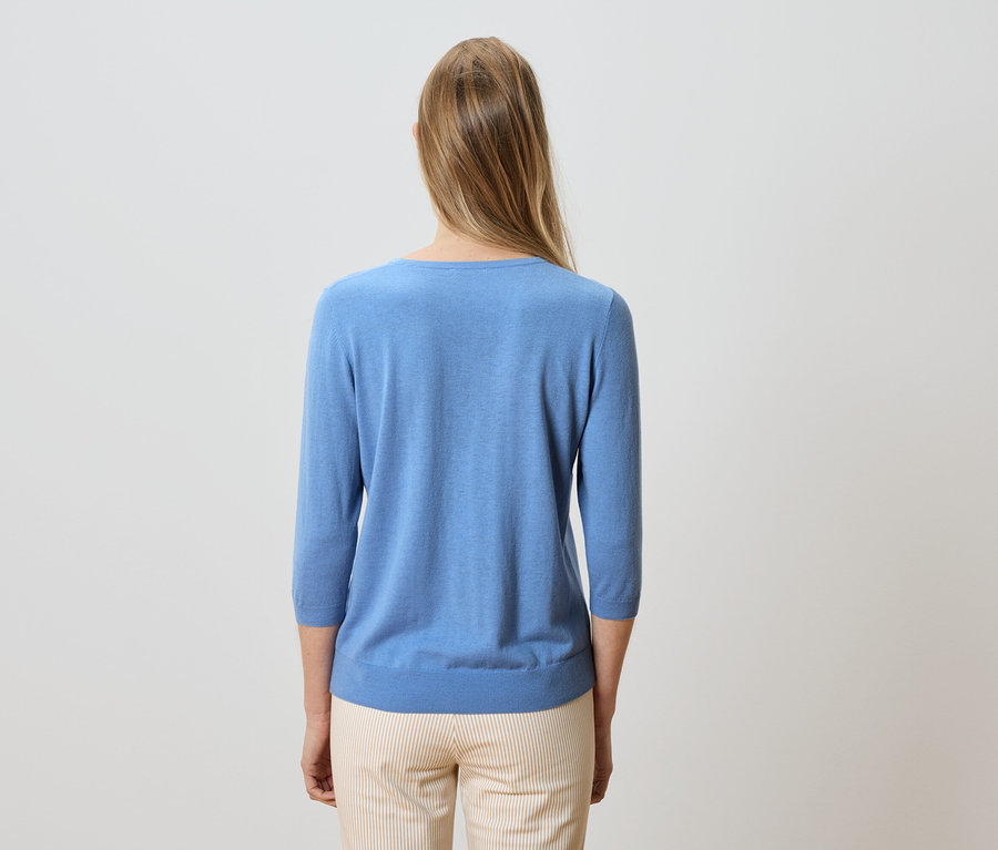 Pohled zezadu na ženu s blond, dlouhými vlasy, oblečenou ve světle modrém svetru a pruhovaných kalhotách.