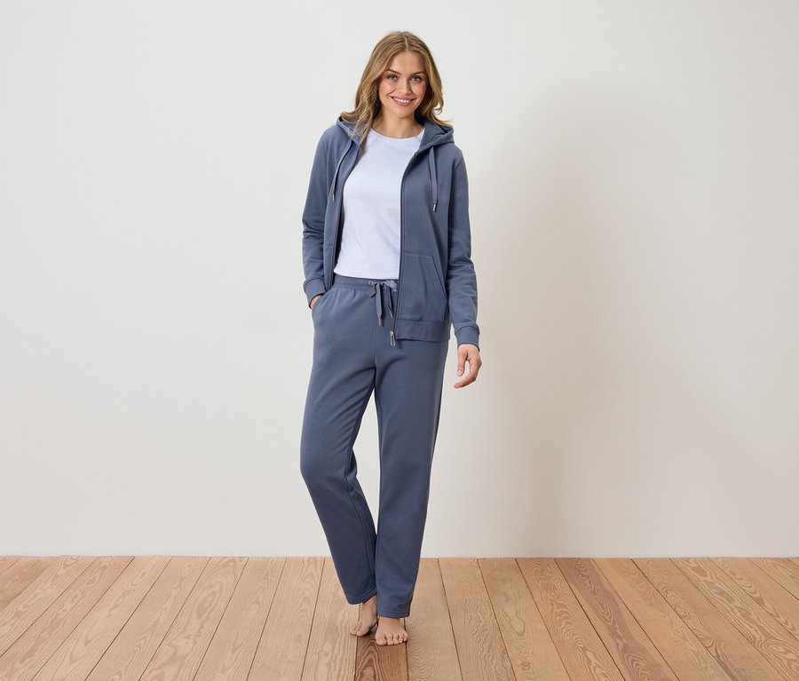 Žena stojí bosá na dřevěné podlaze a má na sobě modrou teplákovou soupravu s mikinou s kapucí a bílým tričkem.