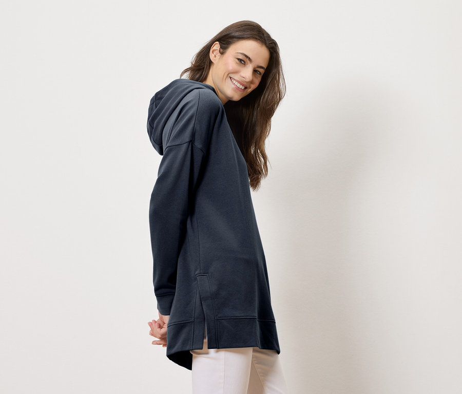 Žena má na sobě dlouhou, tmavě modrou mikinu s kapucí a světlé kalhoty, usmívá se do kamery.