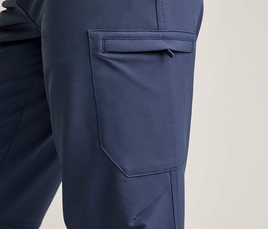 Detail tmavě modrých kalhot s boční cargo kapsou, která má malý zip s poutkem. Látka je hladká s viditelnými švy.