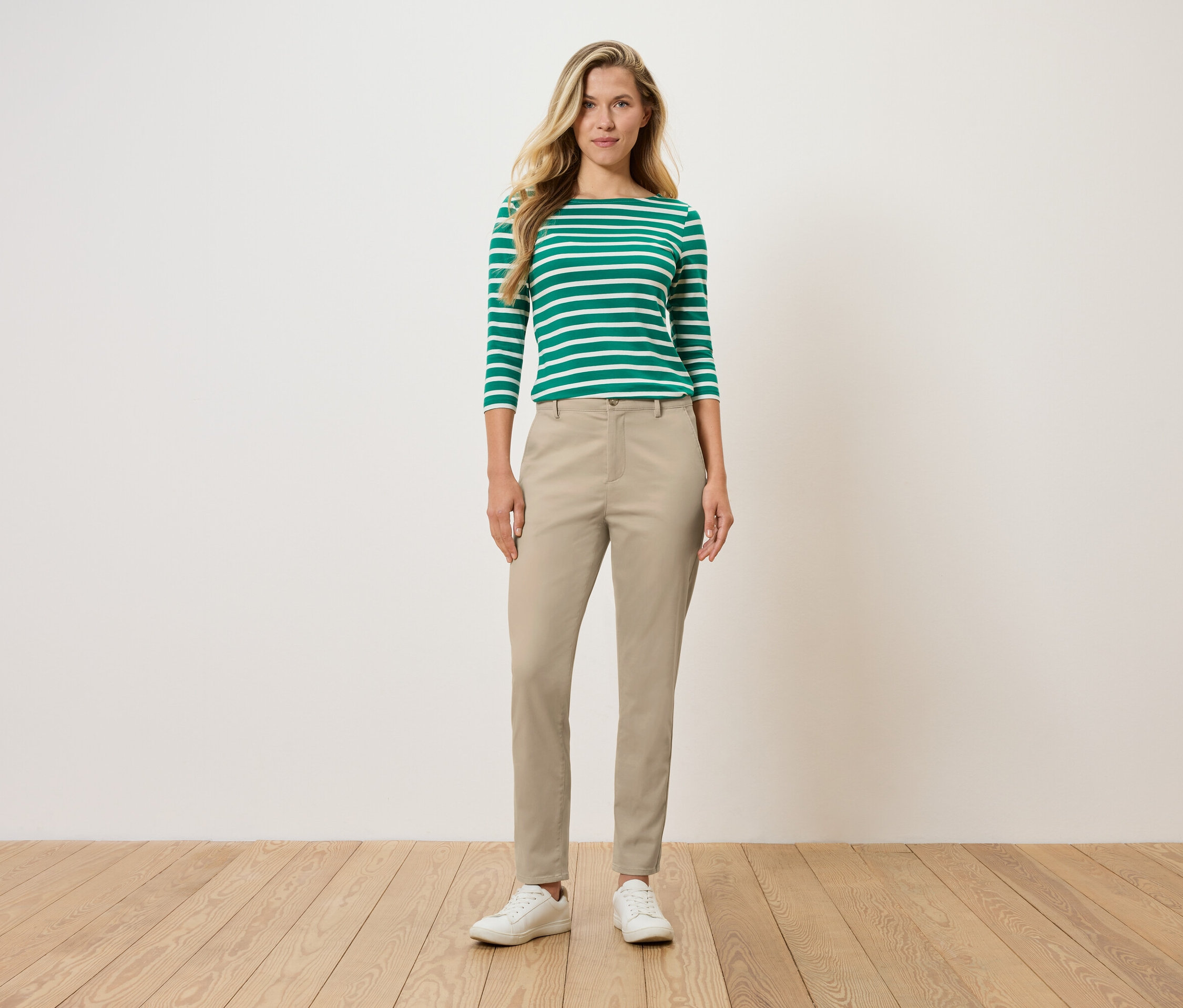 Žena stojí a má na sobě zeleno-bílé pruhované tričko s dlouhým rukávem, béžové kalhoty a bílé boty.