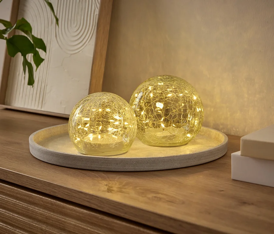 Dvě svítící kulové dekorativní lampy s efektem prasklého skla, naplněné malými LED diodami, stojí na kulatém podnosu na dřevěné komodě. V pozadí je rostlina a zarámovaný obrázek.