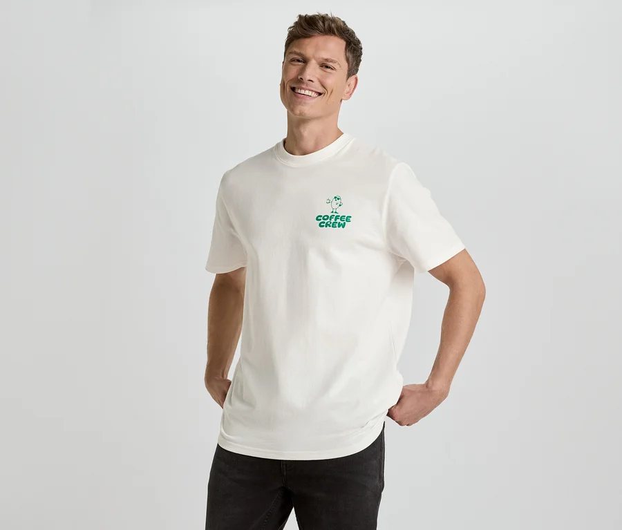 Muž se usmívá a pózuje v krémovém tričku se zeleným potiskem "Coffee Crew" a černých džínách na bílém pozadí.