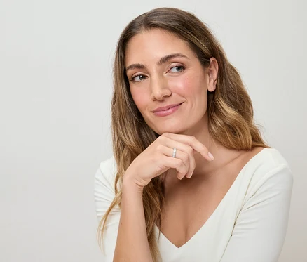 Žena s dlouhými vlnitými vlasy a bílým topem má na prstě stříbrný prsten a zamyšleně se dívá stranou.
