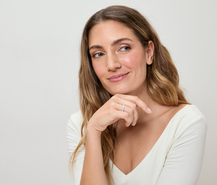 Žena s dlouhými vlnitými vlasy a bílým topem má na prstě stříbrný prsten a zamyšleně se dívá stranou.