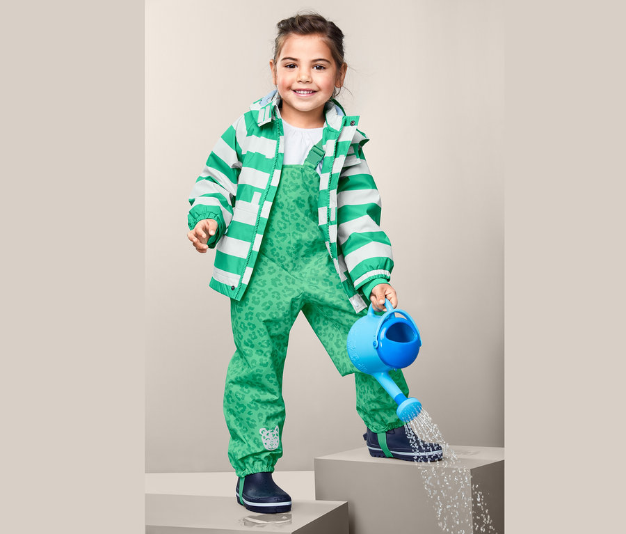 Dívka v zelených kalhotách do deště z recyklovaného materiálu lije vodu z modré konve.