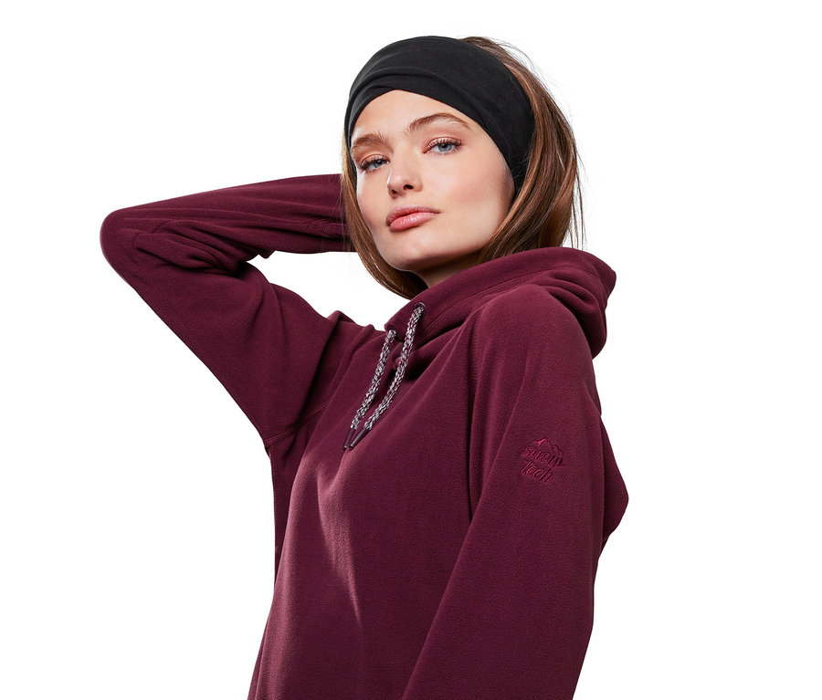Žena s černou multifunkční šálou/čepicí na hlavě a v bordové mikině s kapucí pózuje.