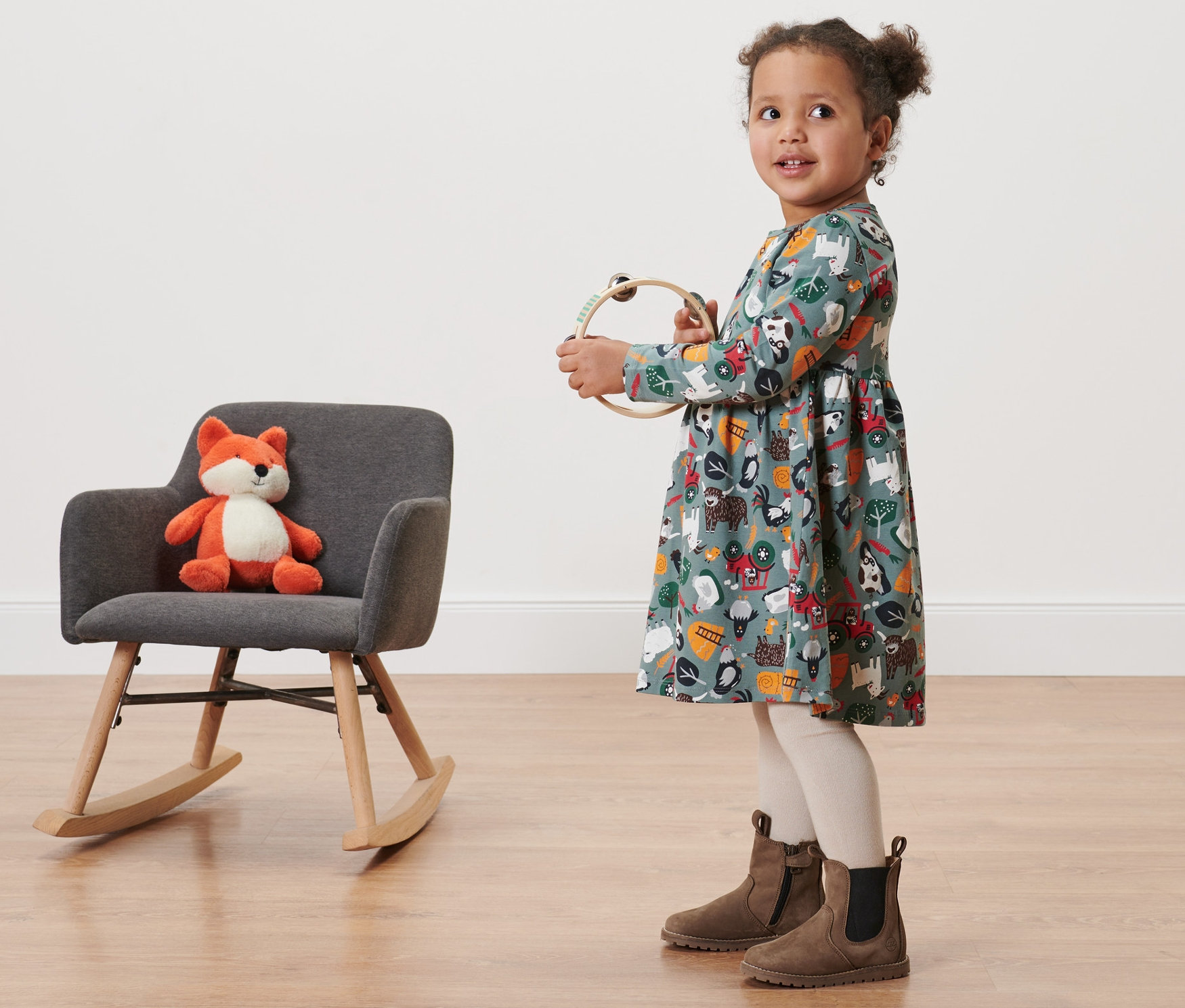 Holčička v žerzejových šatech s potiskem drží tamburínu. Vedle ní stojí šedé houpací křeslo s plyšovou liškou.