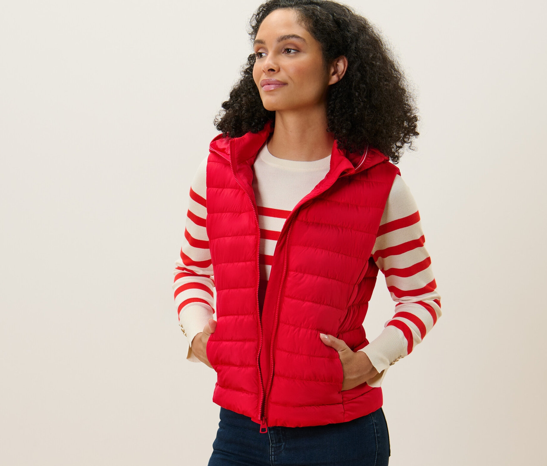 Žena má na sobě prošívanou vestu s kapucí červené barvy. Stojí a dívá se doleva.