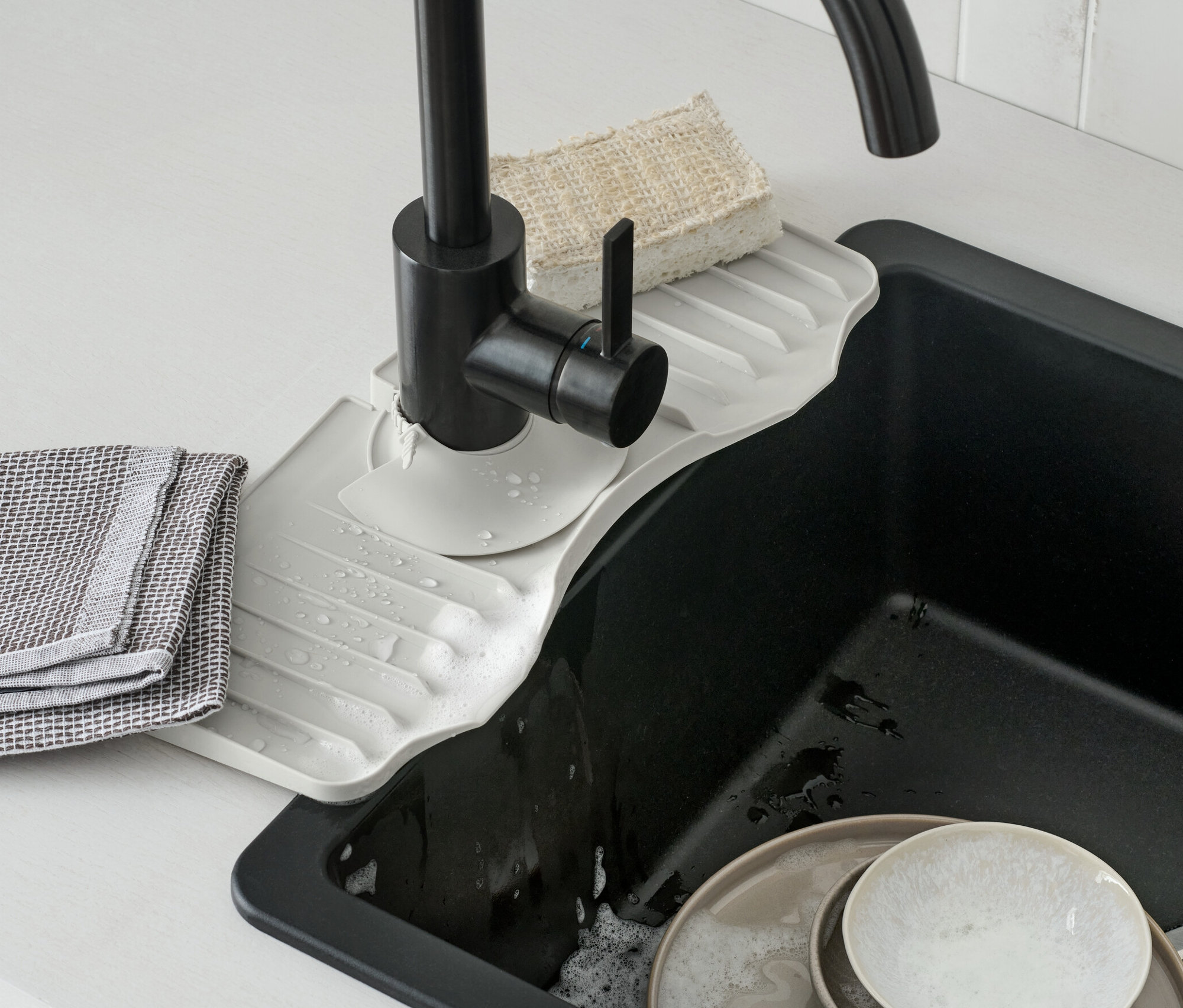 Silikonová podložka pod vodovodní kohoutek je umístěna pod černou vodovodní baterií. V umyvadle se nachází špinavé nádobí.