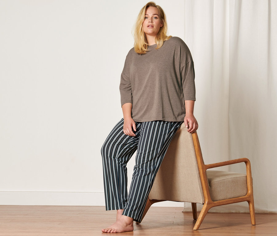 Blonďatá žena pózuje v pyžamu s pruhovanými kalhotami a hnědém topu opřená o křeslo.