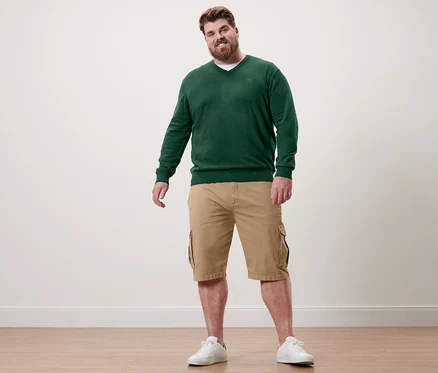 Muž stojí a má na sobě tmavě zelený svetr s výstřihem do V, khaki šortky a bílé tenisky.