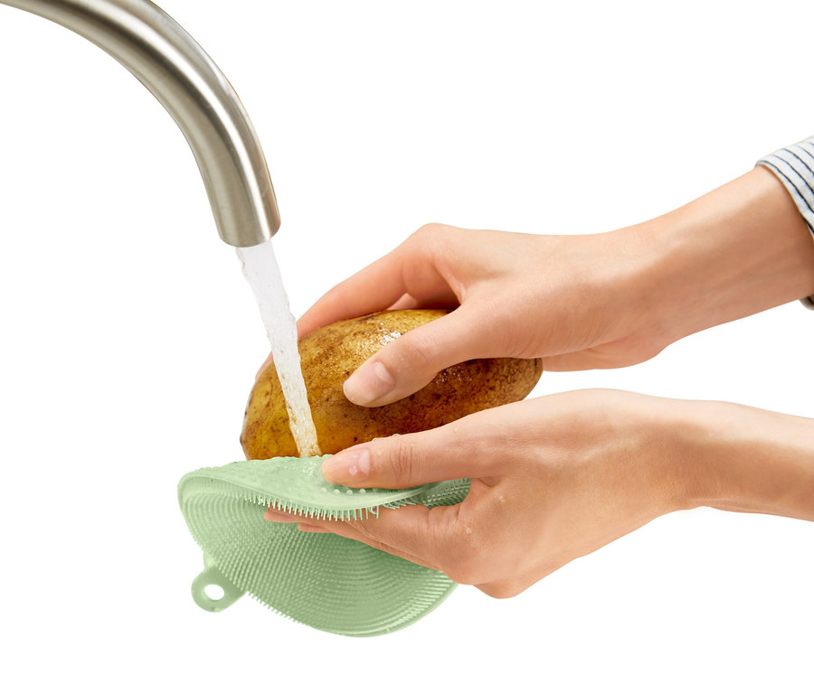Osoba myje bramboru pod tekoucí vodou pomocí silikonové houbičky na nádobí.