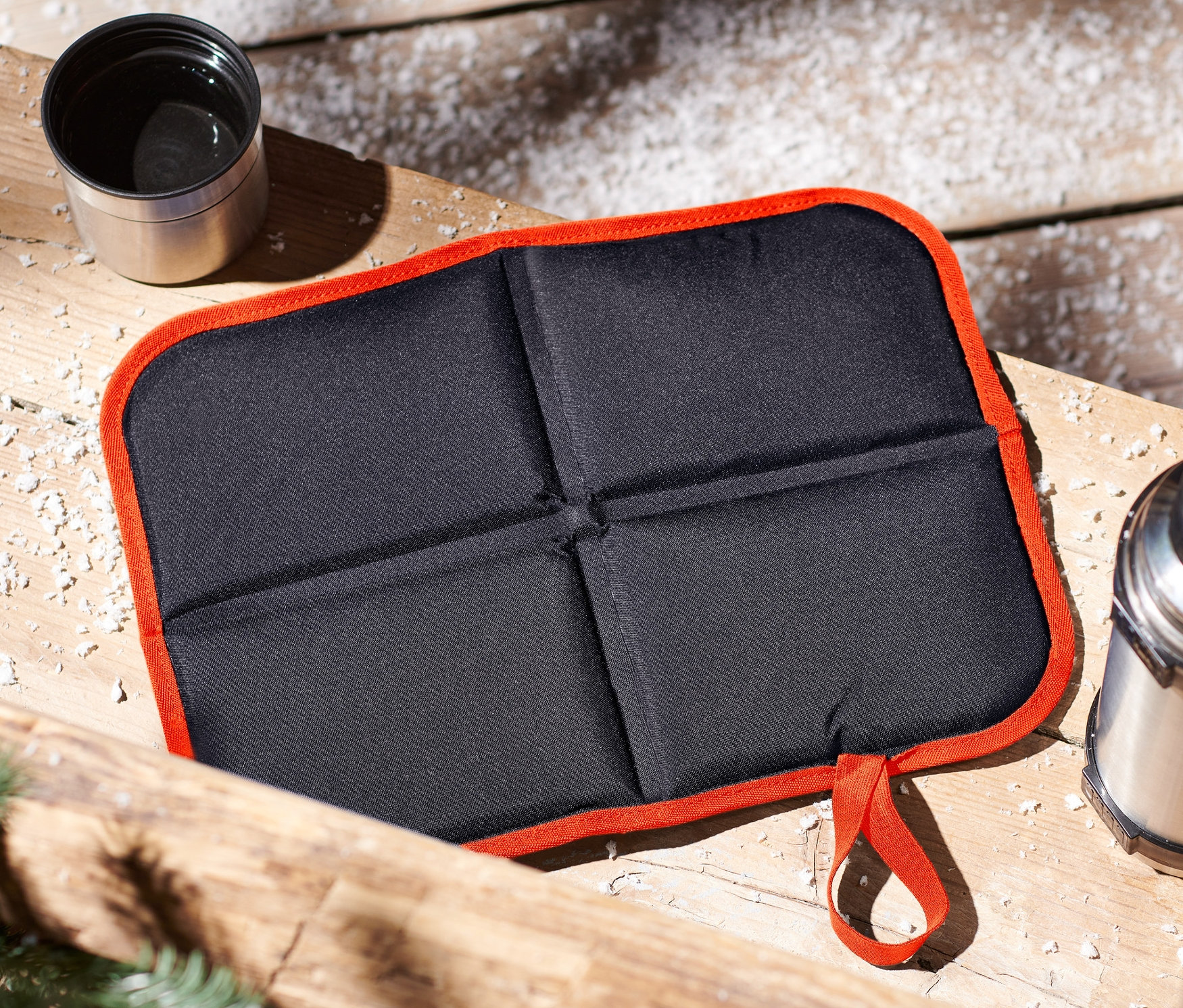Na dřevěném povrchu leží složený černý podsedák s oranžovým lemem, termoska a šálek.