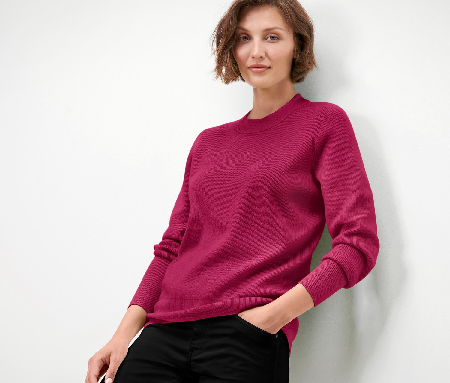 Žena opřená o zeď má na sobě malinově červený svetr z jemného úpletu a černé kalhoty.