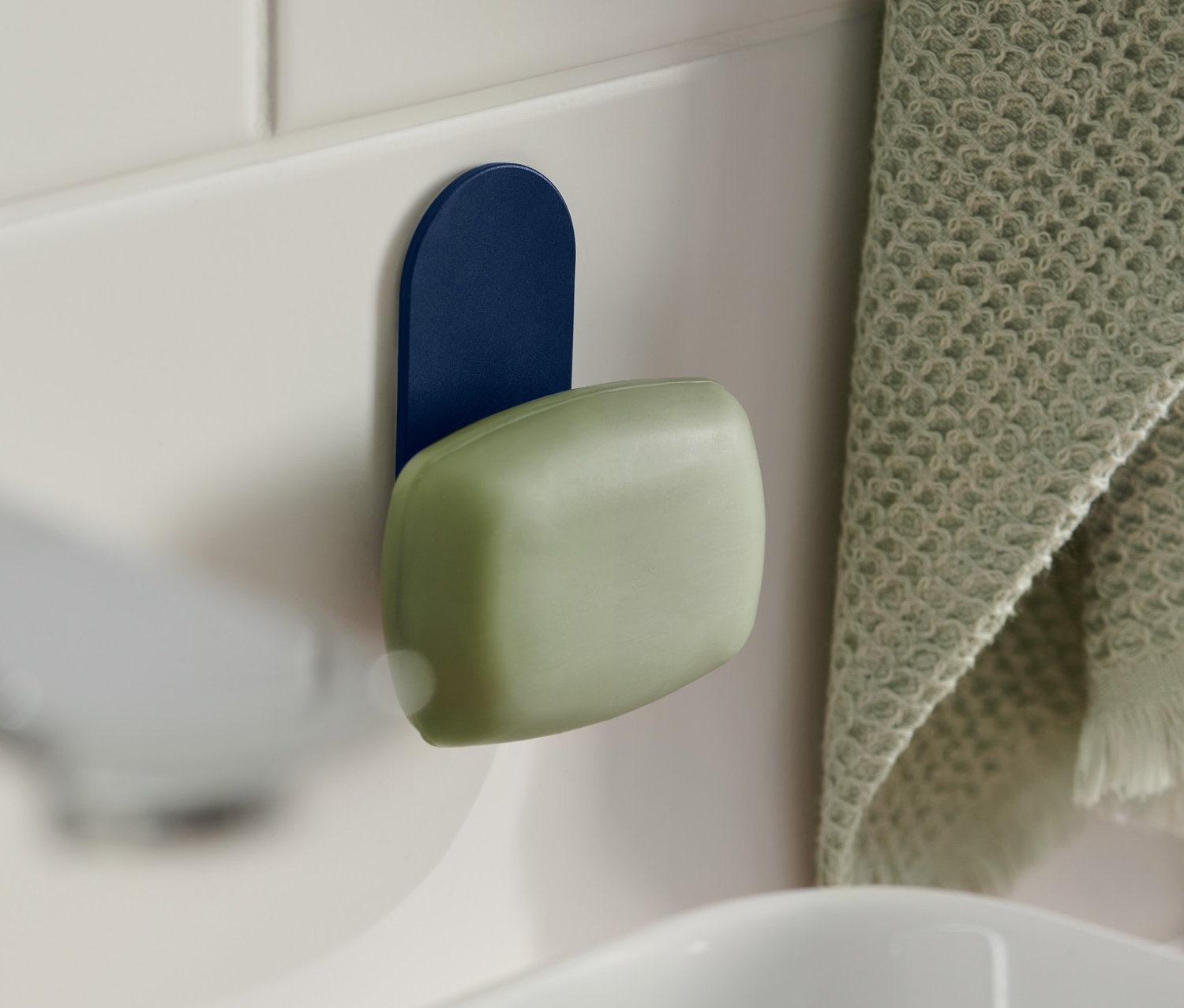 Zelené mýdlo visí na magnetickém držáku na mýdlo modré barvy, který je připevněn na bílých kachličkách u umyvadla. Vpravo visí zelený ručník.