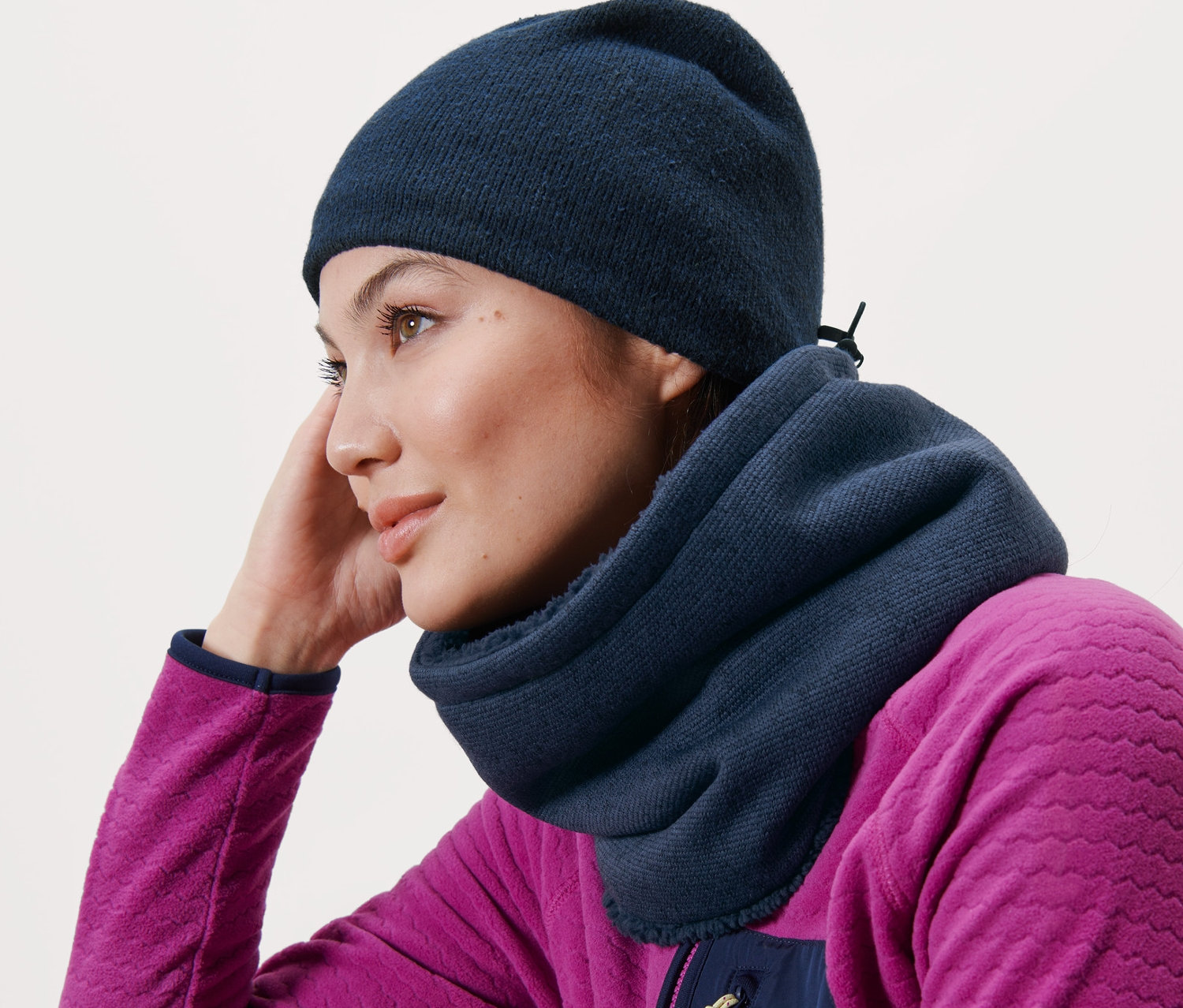 Žena opřená o dřevěnou lavici má na sobě modrý pletený fleecový kruhový šál a modrou čepici.