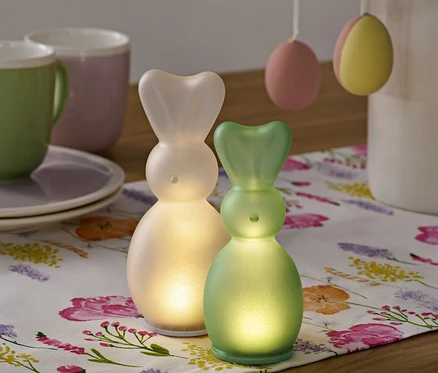 Dva LED zajíčci stojí na květinovém běhounu, v pozadí jsou šálky a velikonoční vajíčka.