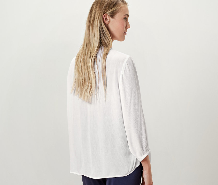 Blondýna pózuje zády ke kameře a má na sobě bílou tunikovou halenku.