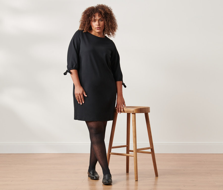 Žena stojí a pózuje vedle dřevěné stoličky, má na sobě černé žerzejové šaty a puntíkované punčocháče.