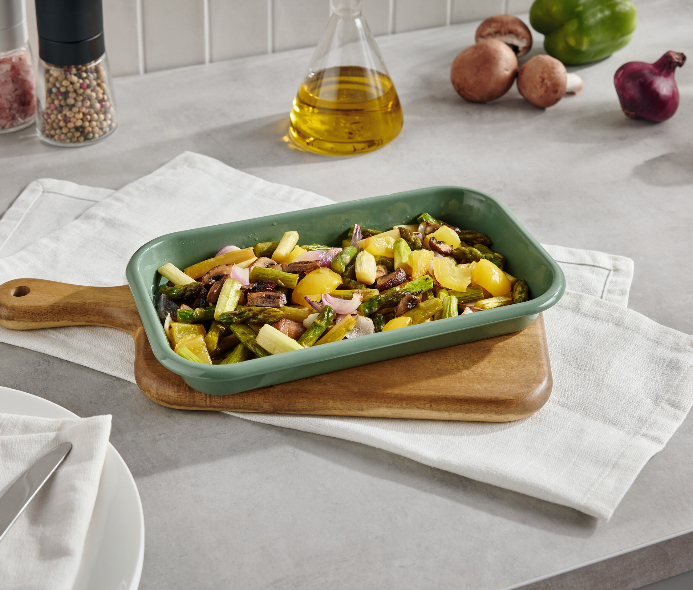 Zelený pekáč s upečenou zeleninou na prkénku s ubrouskem. V pozadí jsou houby, paprika, cibule a kořenky.