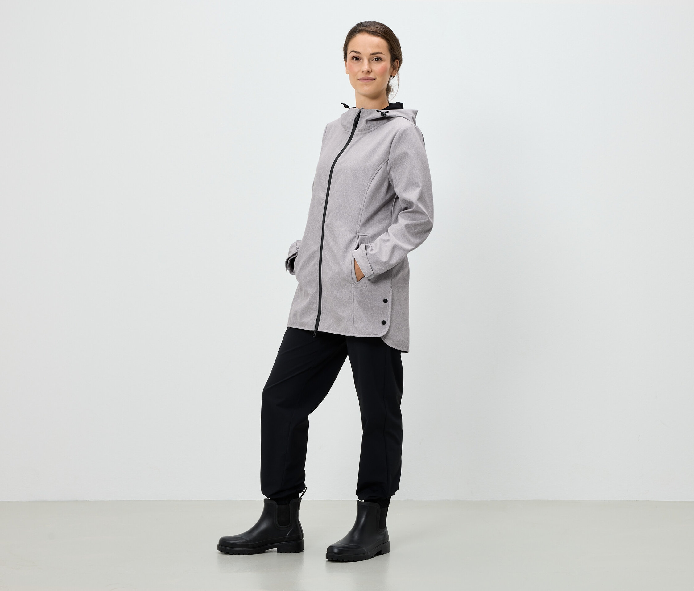 Žena stojí a má ruce v kapsách šedé bundy s kapucí, černé kalhoty a černé boty.