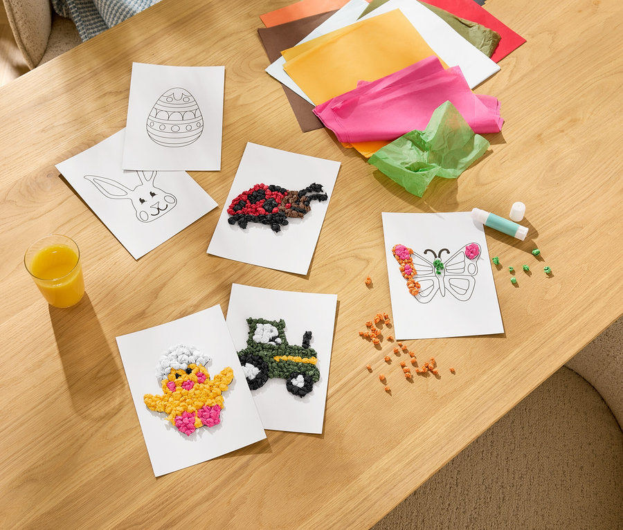 Na dřevěném stole leží ruční práce, včetně lepených kartiček s kuřátkem, traktorem, motýlem a želvou. Vidět je také barevný hedvábný papír, tyčinkové lepidlo a sklenice džusu.