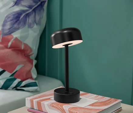 Černá bateriová stolní lampa s LED na knize s květinovým polštářem v pozadí.