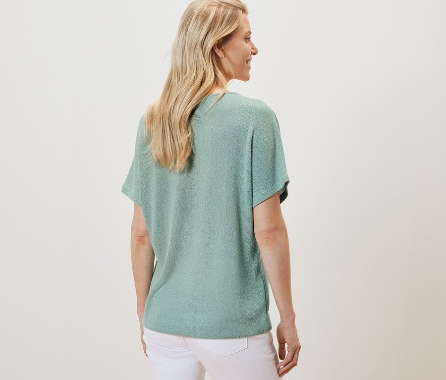 Zezadu je vidět žena s blond vlasy v tričku šalvějové barvy a bílých kalhotách.