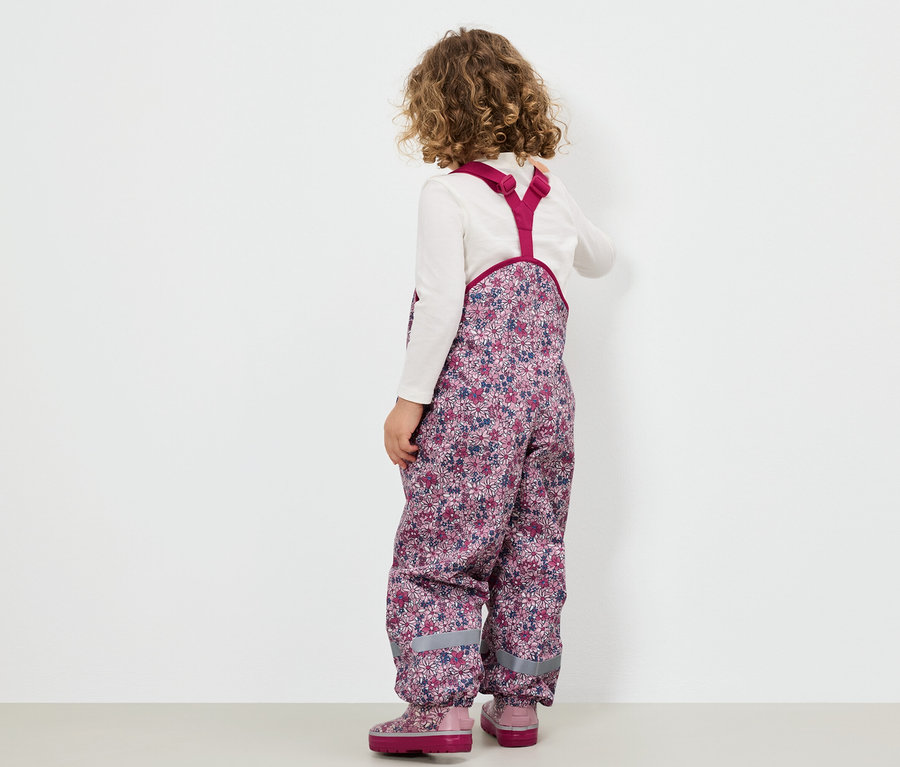 Dívka v květinových kalhotách s laclem a růžových botách stojí zády k divákovi.