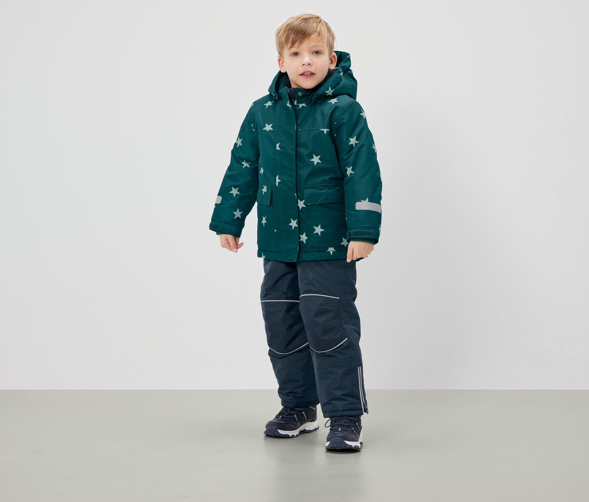 Chlapec stojí v zelené zimní bundě s hvězdami a tmavě modrých kalhotách.
