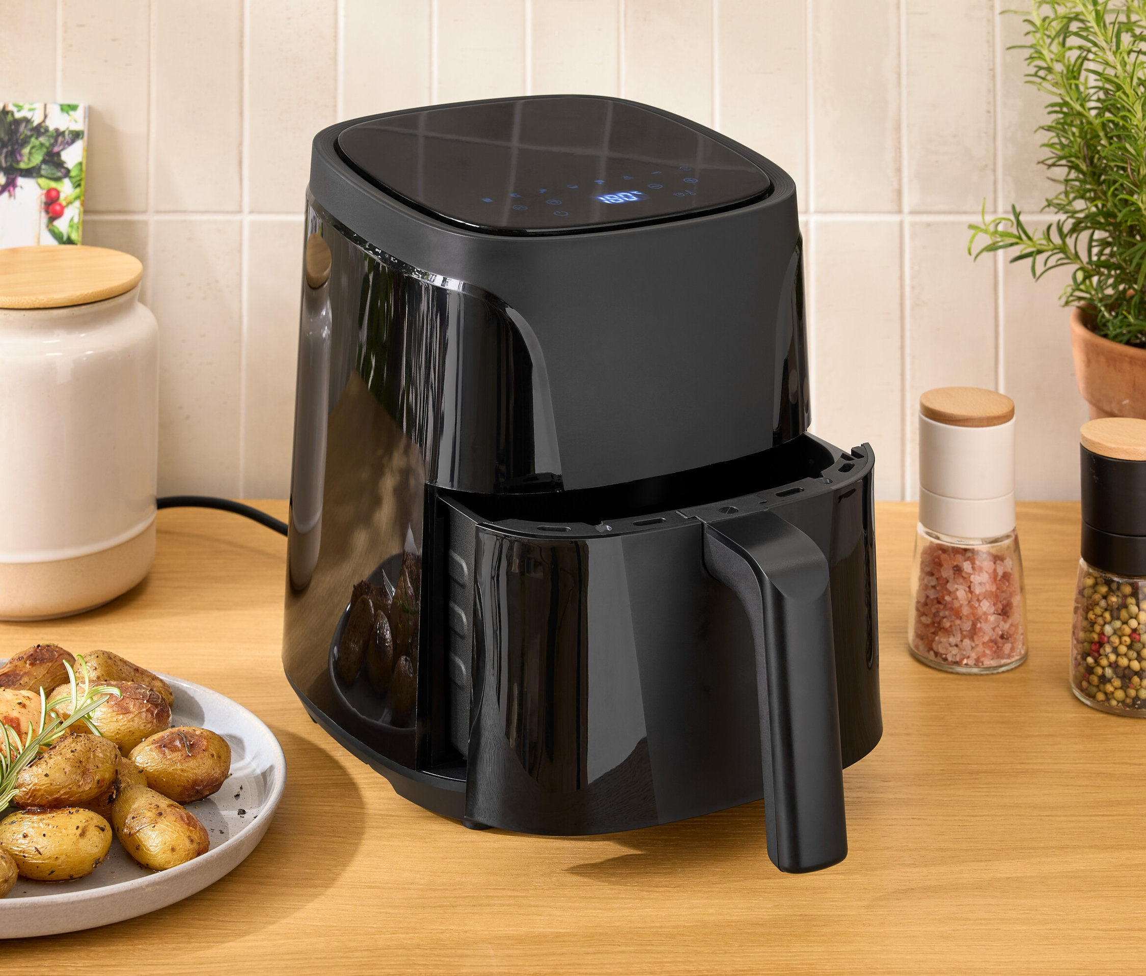 Černá horkovzdušná fritéza na dřevěném stole vedle talíře s kuřetem a bramborami.