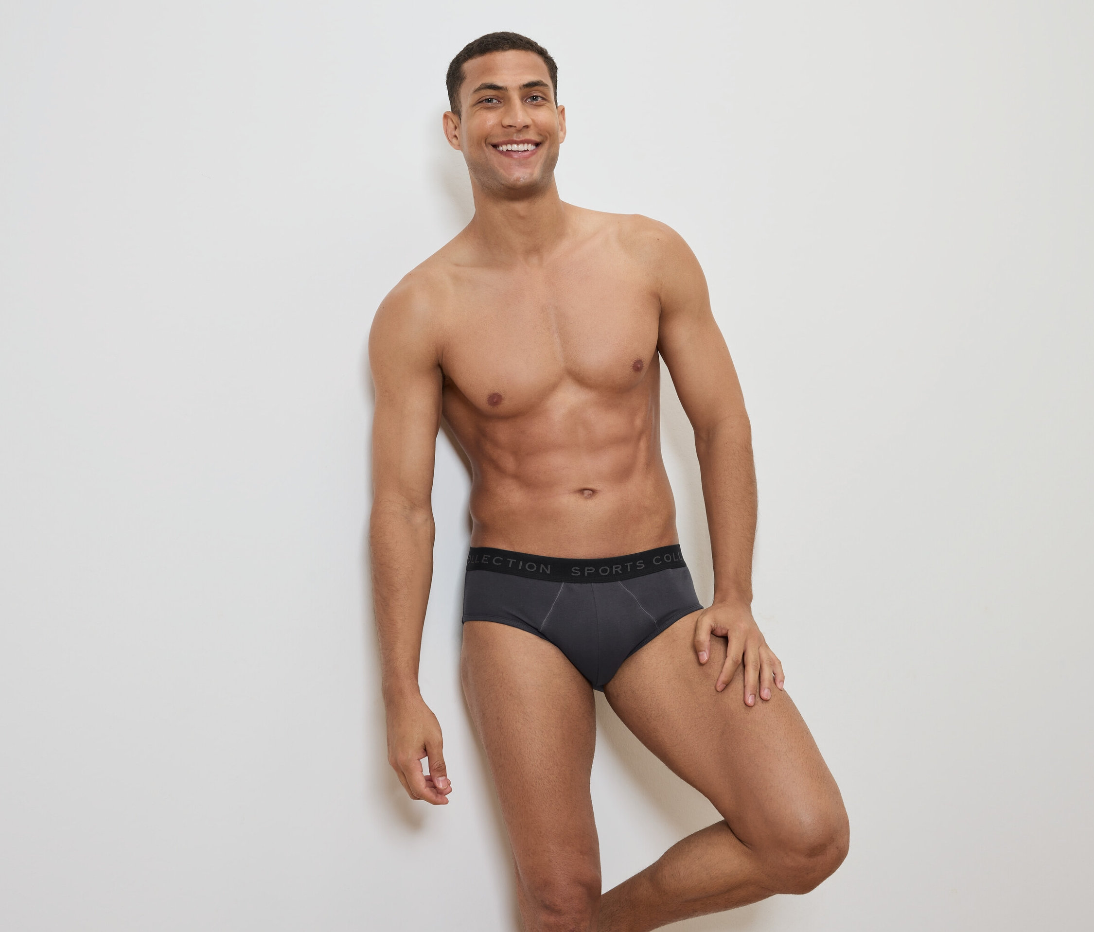 Usmívající se muž s odhalenou horní částí těla pózuje v šedých slipových kalhotkách s černým pasem.