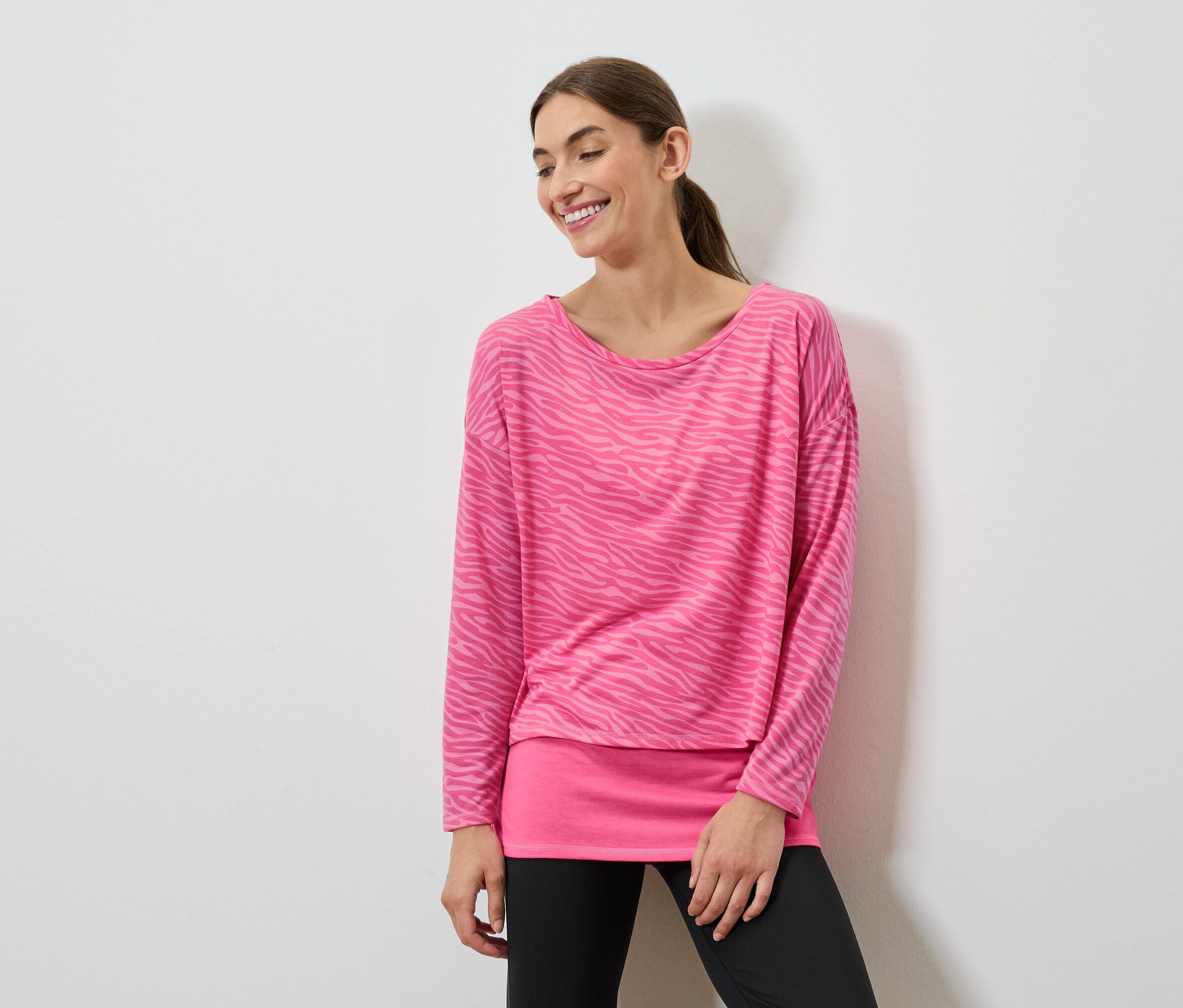 Usmívající se žena pózuje v růžovém topu s dlouhým rukávem a černých legínách.