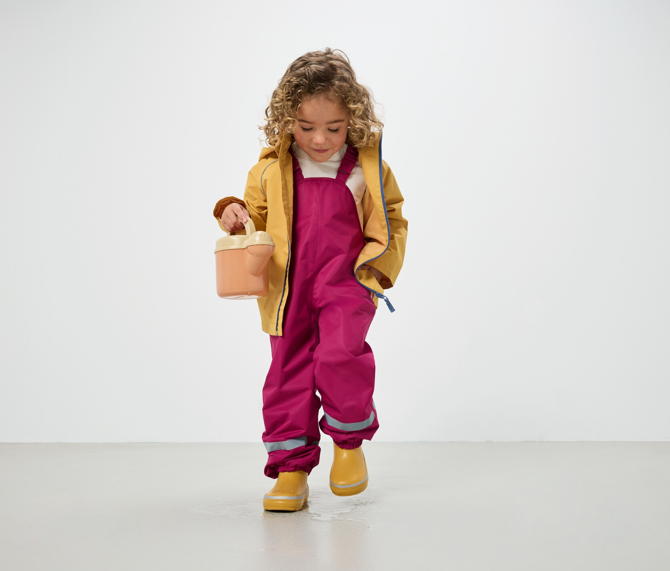 Dítě s kudrnatými vlasy v žluté bundě, růžových kalhotách a žlutých holínkách drží konev a stojí na podlaze.