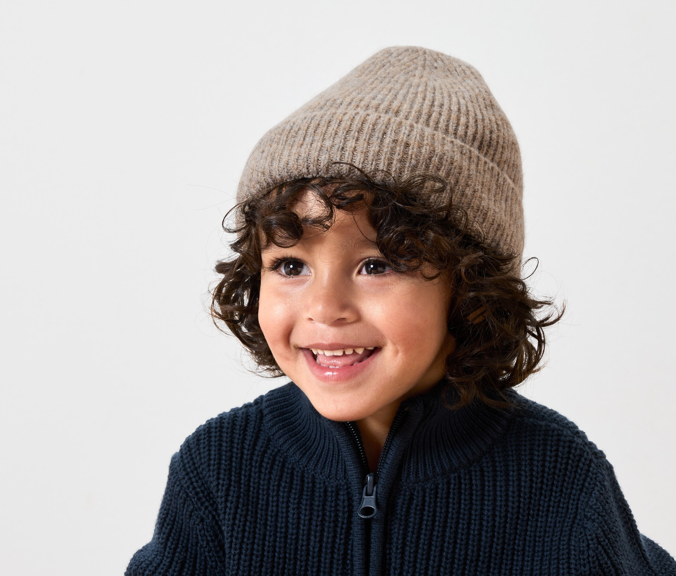 Usmívající se chlapec s hnědými kudrnatými vlasy má na sobě béžovou vlněnou čepici a tmavě modrý pletený svetr na zip.