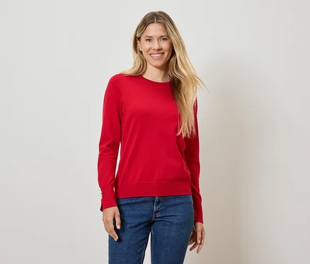 Žena s blond vlasy pózuje v červeném svetru a modrých džínách.