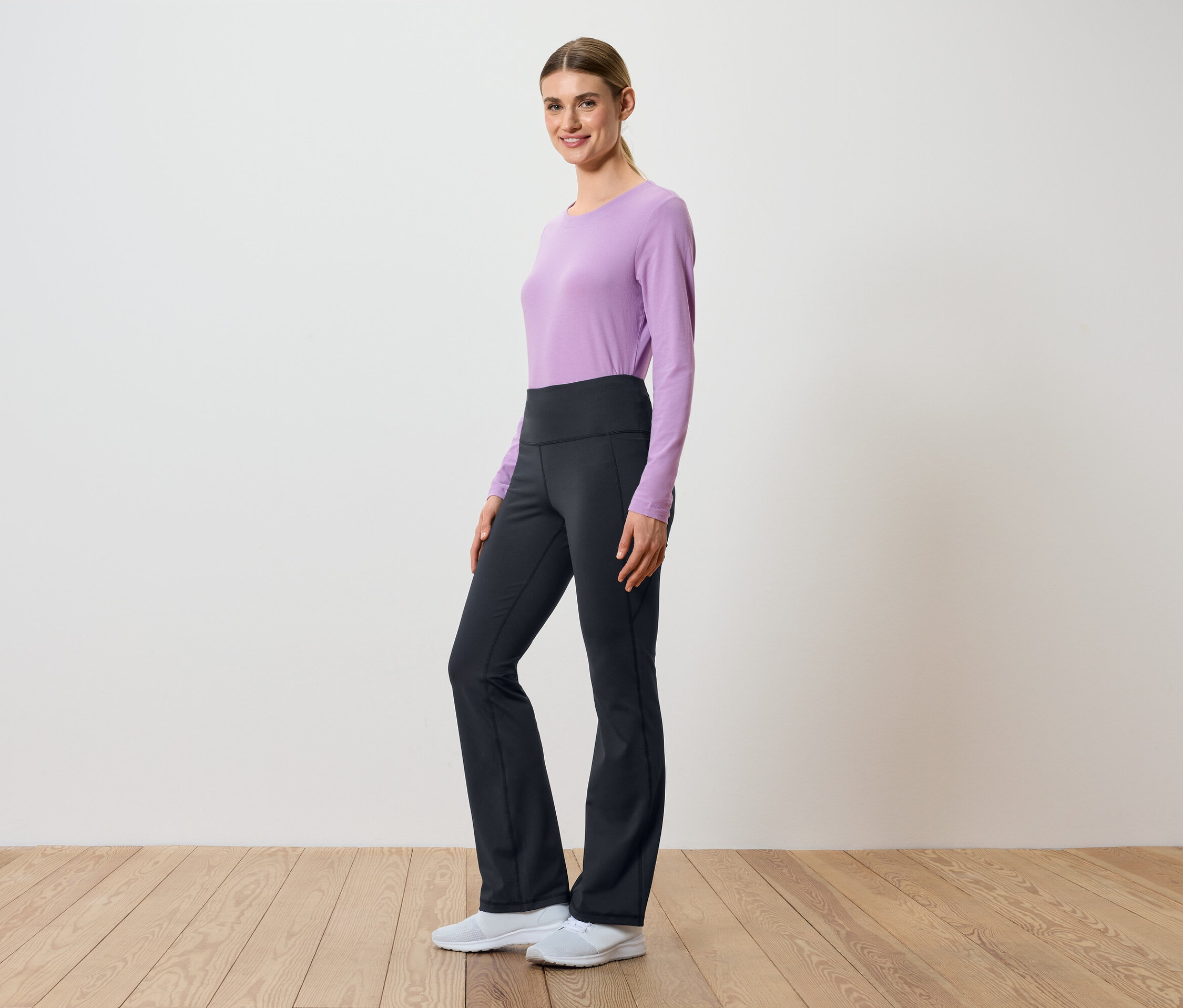 Žena pózuje bokem v lila topu s dlouhým rukávem a černých kalhotách s vysokým pasem.