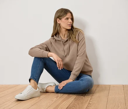 Žena sedí na dřevěné podlaze, oblečená v hnědé mikině s kapucí, modrých džínách a bílých teniskách.