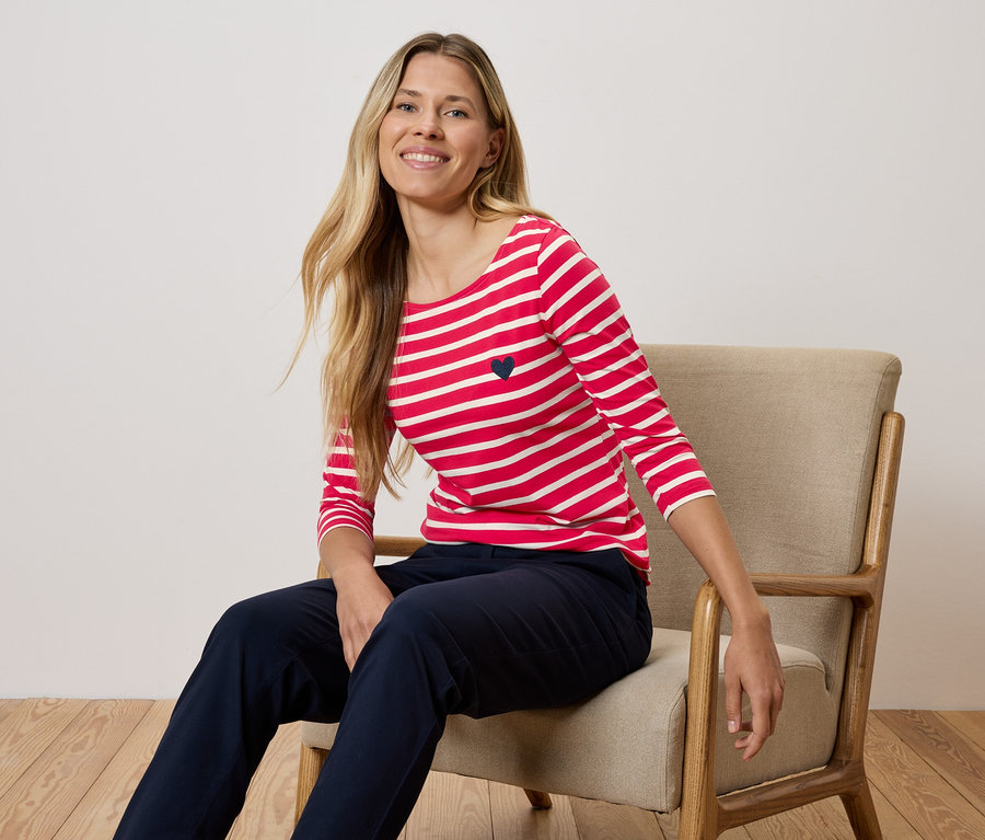 Žena sedí na židli a má na sobě červeno-bílé pruhované tričko a tmavě modré kalhoty.