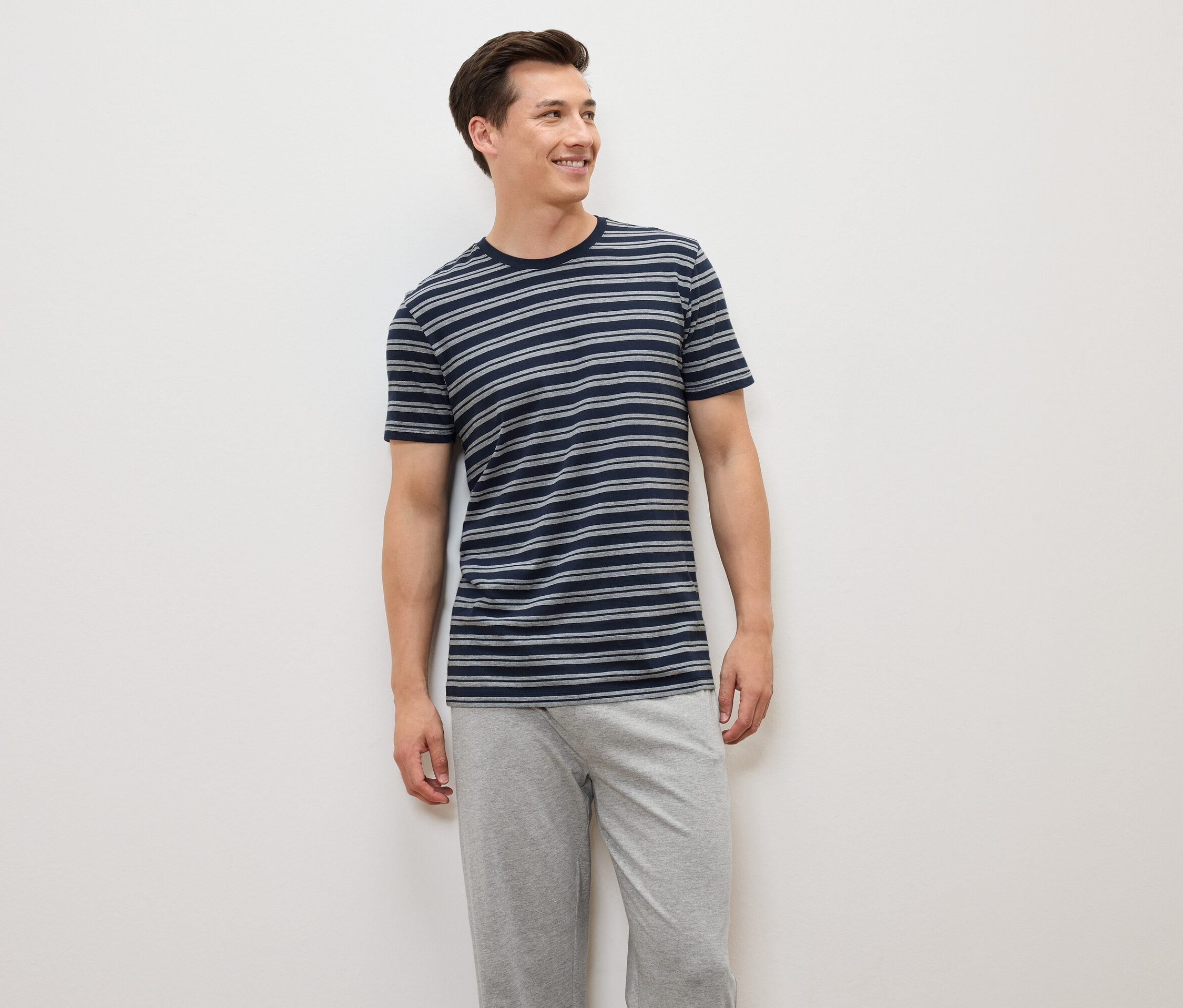 Muž stojí a má na sobě modrošedé pruhované tričko a šedé kalhoty.