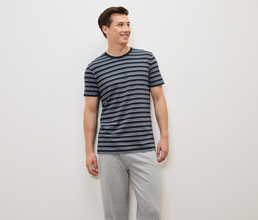 Muž stojí a má na sobě modrošedé pruhované tričko a šedé kalhoty.