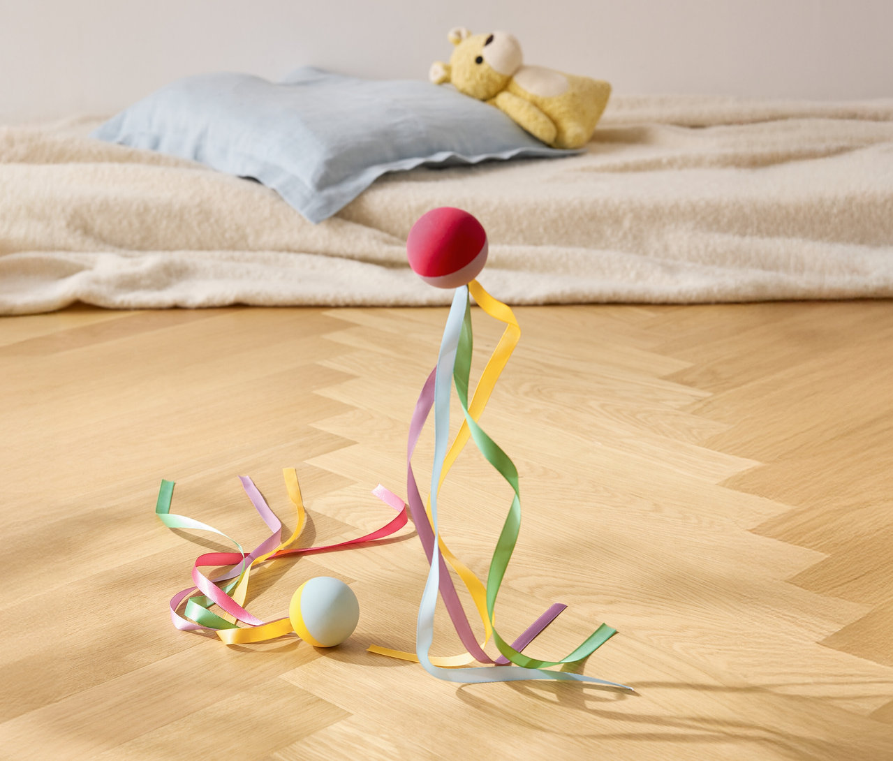 Červený míč stojí s barevnými stuhami na dřevěné podlaze, v pozadí polštář a plyšový medvídek.